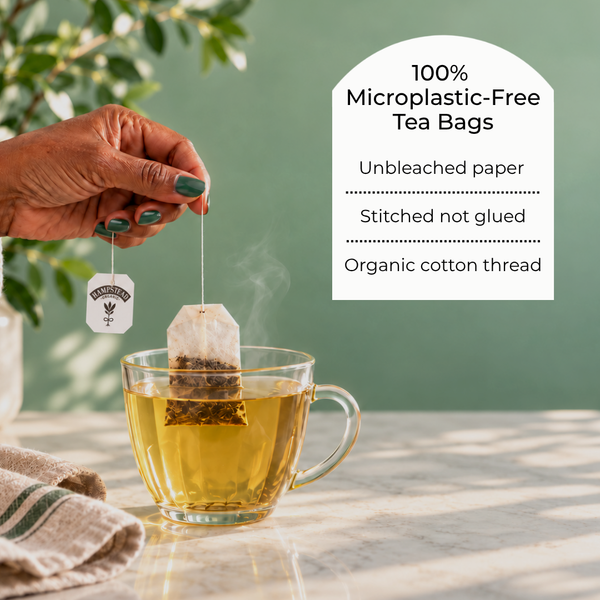 A hand with teal nail polish holds a tea bag above a glass teacup on a marble surface, with a green wall and leafy plant in the background. A white text panel on the right reads '100% Microplastic-Free Tea Bags' with bullet points about unbleached paper, stitched not glued, and organic cotton thread.